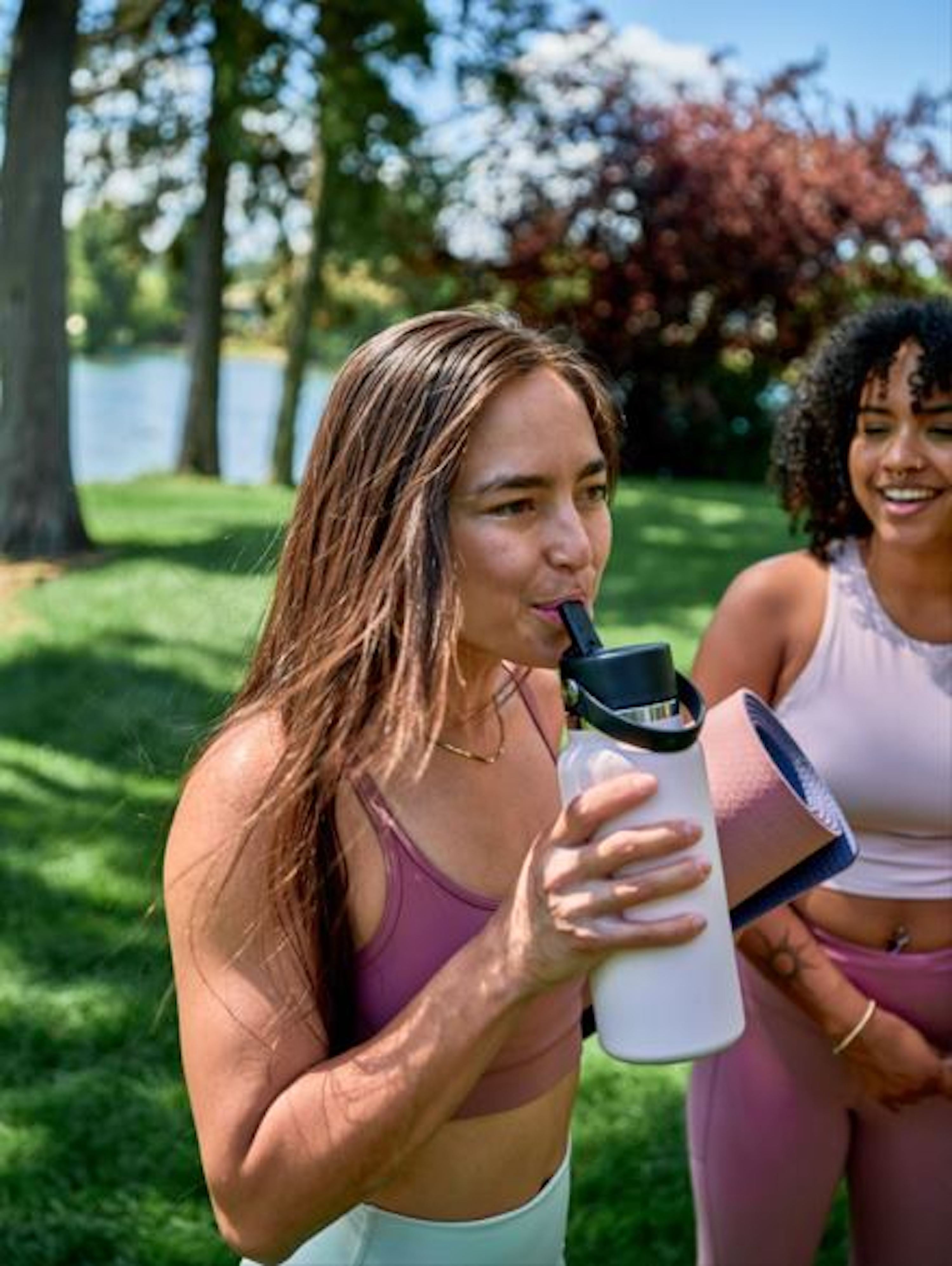 Two women in athletic wear standing outdoors by a lake, one holding a water bottle.